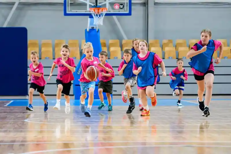 Children in pink and blue training bibs running after the ball in a basketball hall
